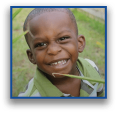 A child wearing a green and white striped shirt is smiling outdoors with a blade of grass near their face. Description generated by AI