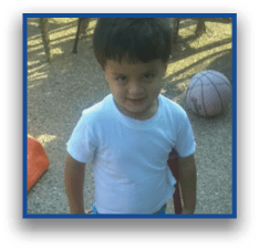 A child with short hair wearing a white shirt and colorful shorts stands outdoors near a small orange slide, a basketball, and patio furniture. Description generated by AI