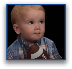 A child wearing a plaid shirt is holding a small football and looking slightly upward against a dark background. Description generated by AI