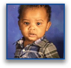 A child with short curly hair wearing a plaid button-up shirt looks directly at the camera against a blue background. Description generated by AI