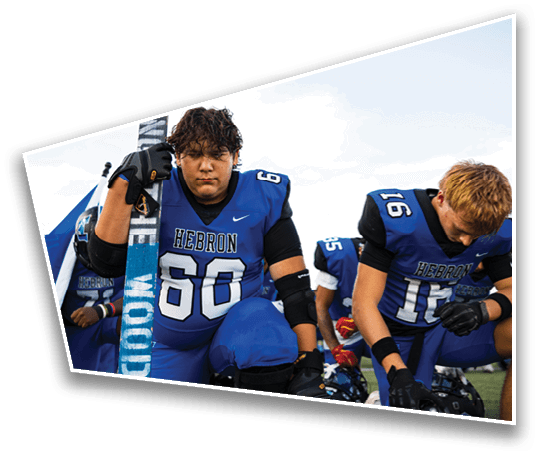 Two people in blue football uniforms kneel on a field, one holding a flagpole with the word “WOOD" visible. Description generated by AI