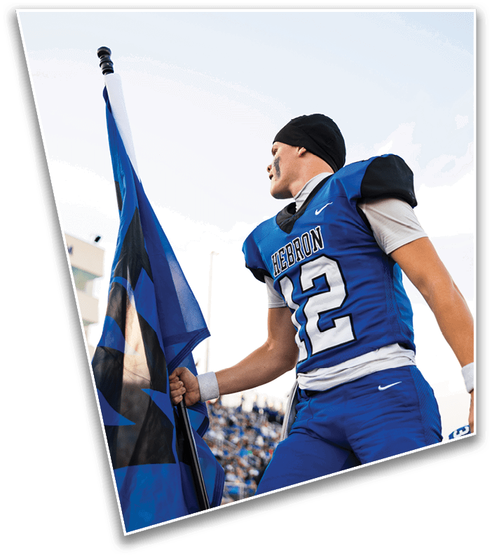 A person wearing a blue football uniform with the number 12 and the word Hebron is holding a flag and looking to the side on a sports field. Description generated by AI