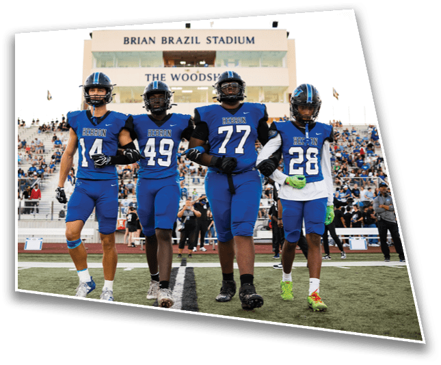 Four people in blue football uniforms walk arm in arm on a field with a stadium and crowd in the background. Description generated by AI