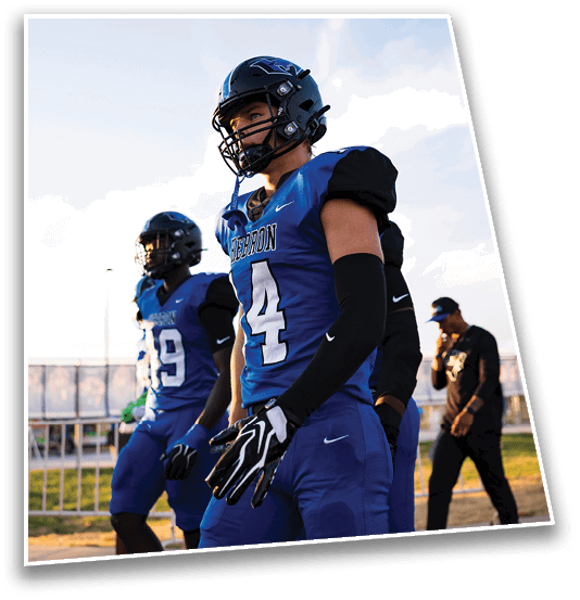 Two people in blue football uniforms and helmets stand outdoors near a fence with a clear sky in the background. Description generated by AI