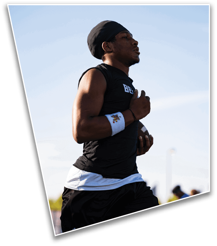 A person wearing athletic clothing and a headband is holding a football and appears to be running or preparing to throw it outdoors with a few blurred people in the background. Description generated by AI