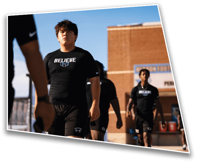 A person wearing a black shirt with the word “BELIEVE" printed on it stands outdoors with two other people partially visible in the background. Description generated by AI