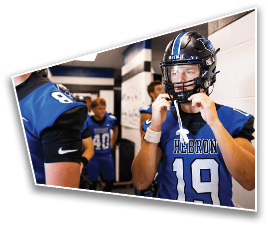 People in blue and black football uniforms are standing in a locker room, with one person adjusting their helmet. Description generated by AI