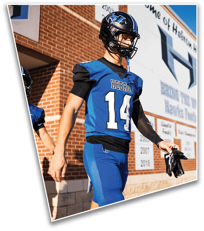 A person wearing a blue football uniform with the number 14 and the word Hebron is walking outside near a brick building while holding a pair of gloves. Description generated by AI