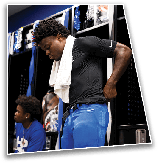 A person wearing a black Nike shirt and blue shorts with a white towel draped around their neck is adjusting their clothing in a locker room. Description generated by AI