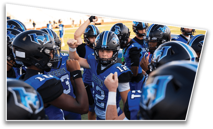 A group of football players in blue and black uniforms and helmets are gathered closely on a field, with one player in the center raising a fist as if giving a motivational speech or cheer. Description generated by AI