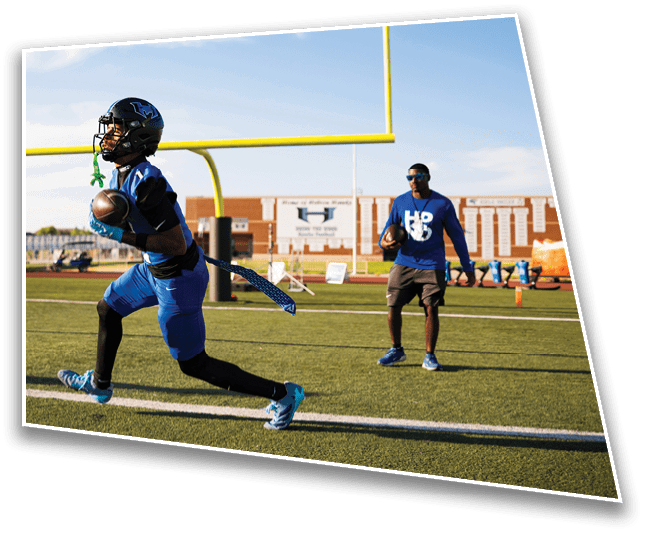A person in blue sports gear is running with a football near the goalpost on a football field while another person in casual athletic clothing watches nearby. Description generated by AI