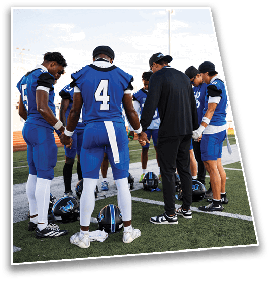 A group of football players and a coach stand in a circle on a field holding hands with helmets placed on the ground inside the circle. Description generated by AI