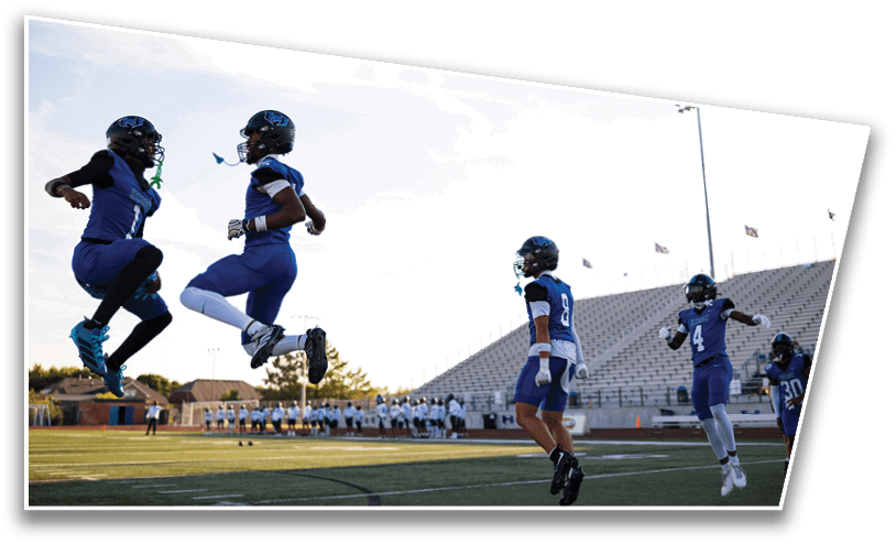 Two people in blue football uniforms are jumping in the air on a football field while two others in similar uniforms stand nearby with a stadium and clear sky in the background. Description generated by AI