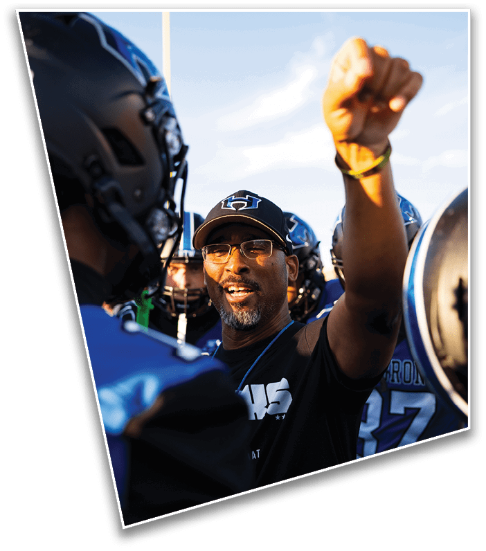 A person wearing glasses and a cap is raising a fist and speaking energetically to a group of people in football helmets and uniforms. Description generated by AI