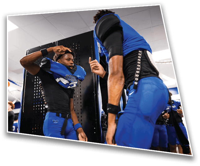 People in blue and black football uniforms are standing in a locker room, with one person appearing to be adjusting their helmet. Description generated by AI