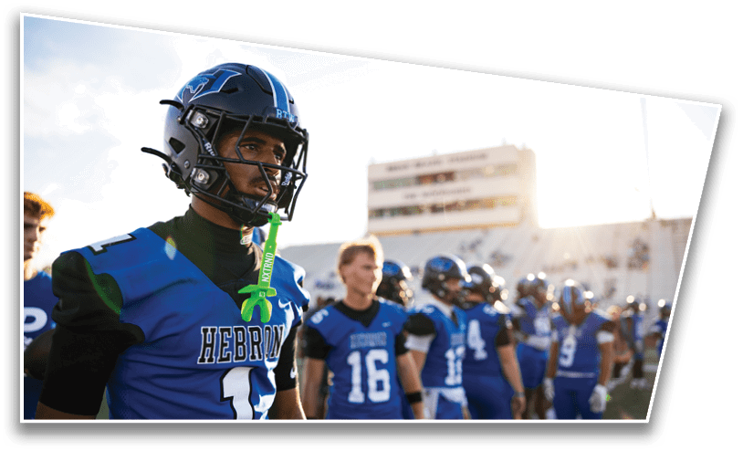 A group of people wearing blue football uniforms and helmets stand on a field with one person in the foreground looking focused. Description generated by AI