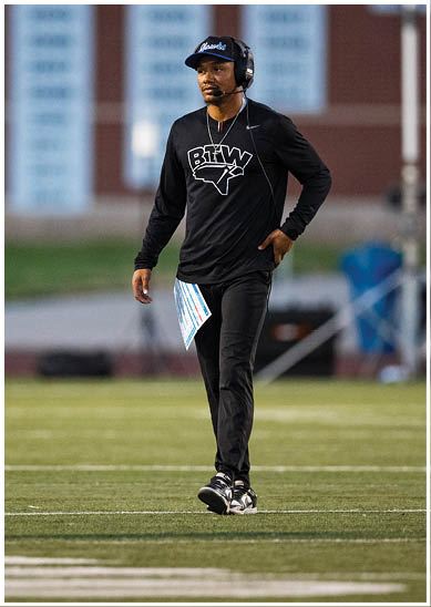A person wearing a headset, black long-sleeve shirt with a sports logo, black pants, and sneakers is walking on a football field holding a play sheet. Description generated by AI