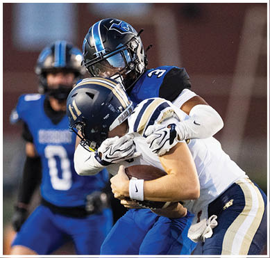 A person in a blue and white football uniform is being tackled by another person in a black and blue football uniform during a game. Description generated by AI