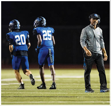 Two people in blue football uniforms with helmets are running on a field while a person in a gray jacket and black pants stands nearby wearing a headset. Description generated by AI