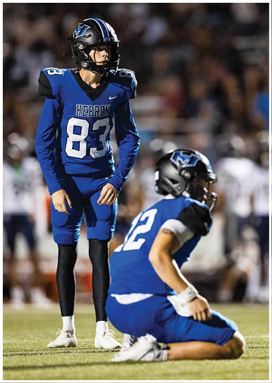 Two people in blue football uniforms are on a field, one kneeling and the other standing, with a blurred crowd in the background. Description generated by AI