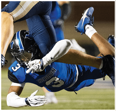 A person in blue sports gear is diving or falling forward on a grassy field while another person in dark pants is partially visible above them. Description generated by AI