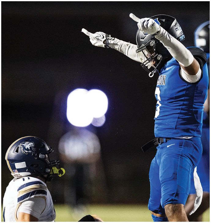 A person wearing a football helmet and gloves is raising both hands with index fingers pointing upward against a dark background. Description generated by AI