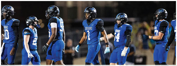 A group of people wearing blue and black football uniforms and helmets are standing on a field, appearing to prepare for a play. Description generated by AI