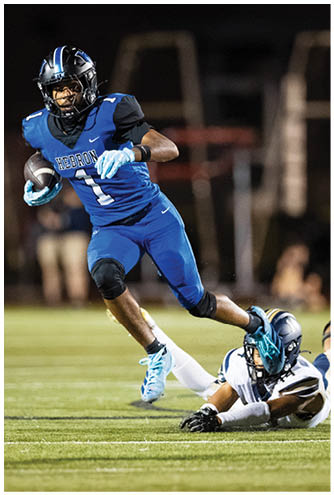 A person in a blue football uniform is running while another person in a white football uniform is on the ground reaching out to grab their leg during a nighttime game. Description generated by AI