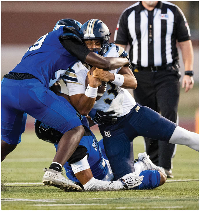 A person in a white and navy football uniform is being tackled by two people in blue football uniforms on a grassy field with a referee standing in the background. Description generated by AI