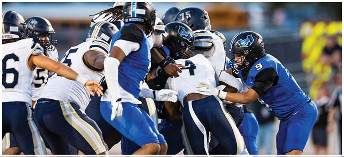 Players in blue and white football uniforms are engaged in a close physical play on a football field during a game. Description generated by AI