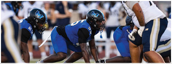 People in football uniforms are crouched on a field, preparing for the start of a play. Description generated by AI