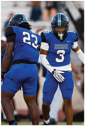 Two people wearing blue football uniforms with helmets are on a field, one facing forward and the other facing away. Description generated by AI