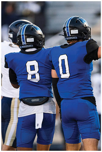Two people in blue football uniforms with helmets are standing side by side on a field, with one raising a fist in celebration. Description generated by AI