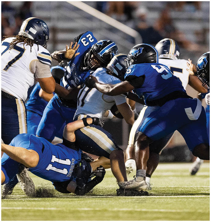 Several football players in blue and white uniforms are engaged in a tackle on a grassy field during a game. Description generated by AI
