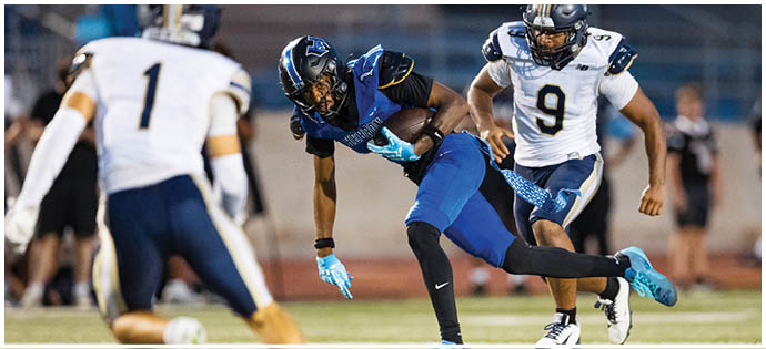 A person in a blue and black football uniform is running with the ball while two people in white and navy uniforms are chasing on a football field. Description generated by AI