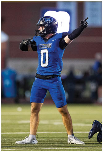 A person in a blue football uniform with the number zero is standing on a field pointing with both hands while two other players in white and blue uniforms are nearby. Description generated by AI