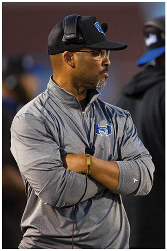 Two adults wearing headsets and casual sportswear stand on the sidelines of a sports field, appearing focused on the game. Description generated by AI