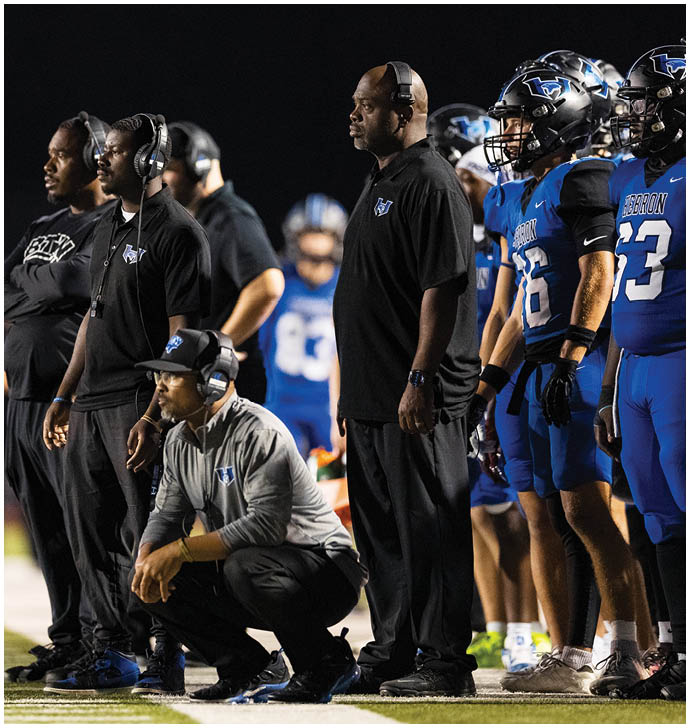 Several people wearing headsets and sports attire stand and crouch attentively on the sidelines of a nighttime football game. Description generated by AI