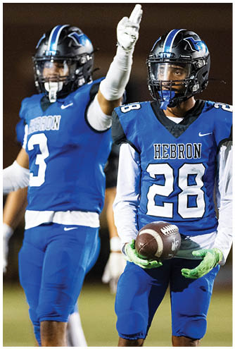 Two people wearing blue football uniforms and helmets are on a field, with one person raising a gloved hand. Description generated by AI