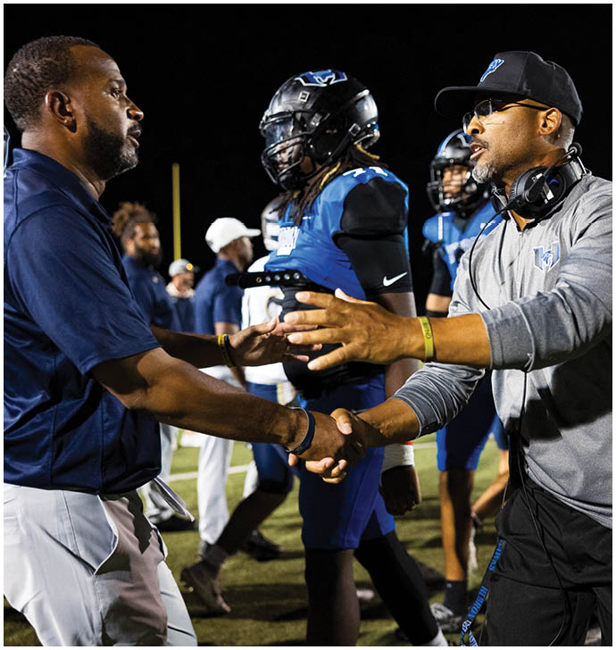 Two adults wearing sports attire shake hands on a football field with players in blue and black uniforms standing in the background. Description generated by AI