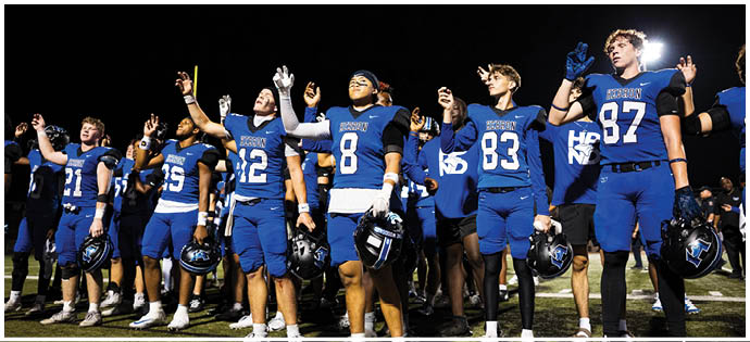A group of people wearing blue football uniforms stand side by side at night with their helmets in one hand and the other hand raised. Description generated by AI