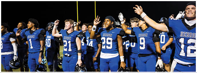 A group of people wearing blue football uniforms stand side by side on a field at night, raising their hands in a celebratory gesture. Description generated by AI
