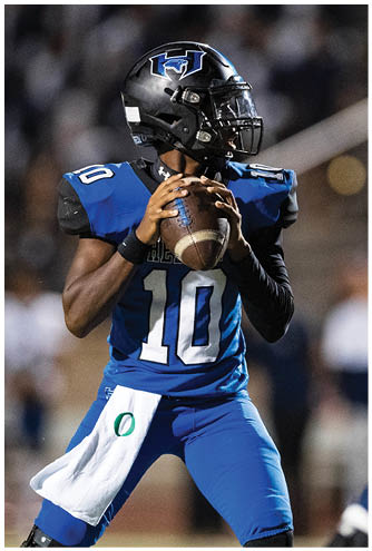 A person wearing a black helmet and blue football uniform is holding a football and preparing to throw it during a game. Description generated by AI