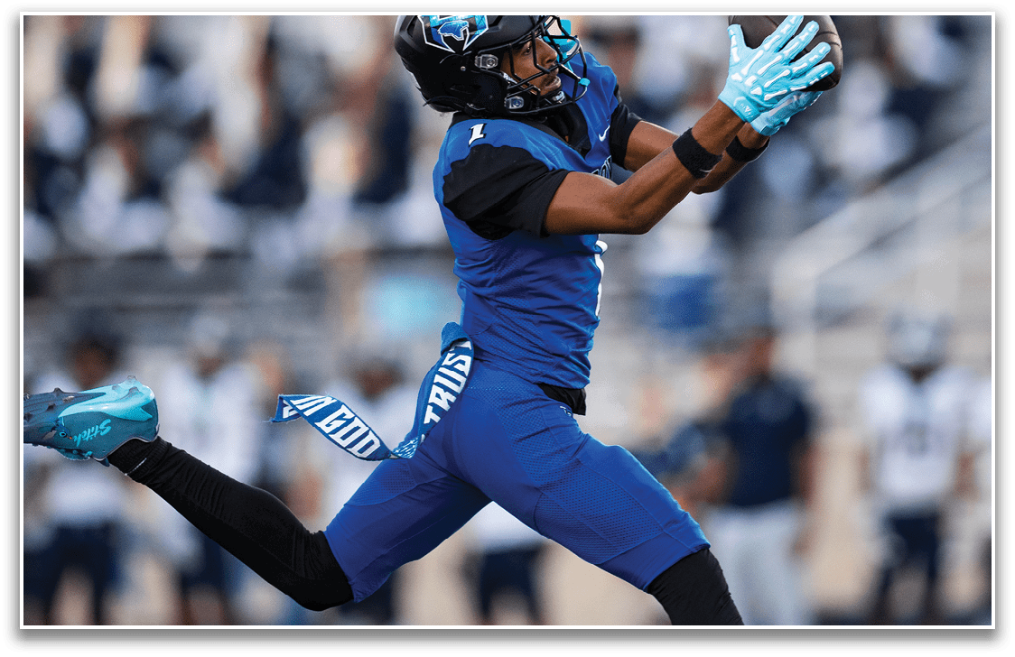 A person wearing a blue sports uniform and gloves is mid-air, reaching out to catch a ball during a football game. Description generated by AI