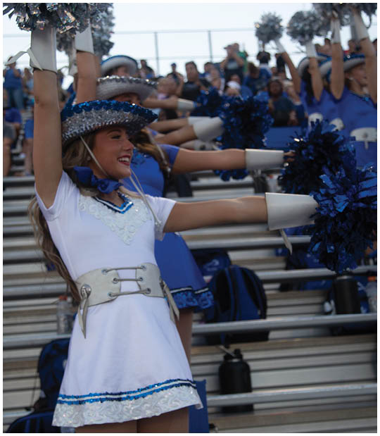 A person wearing a white uniform with a decorative belt and a blue sequined hat is smiling and raising pom-poms while performing in front of a crowd seated on bleachers. Description generated by AI