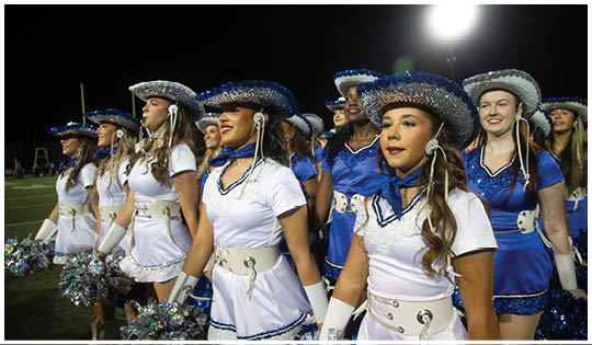 A group of people dressed in coordinated white and blue outfits with decorative hats stand in a line on a sports field at night. Description generated by AI