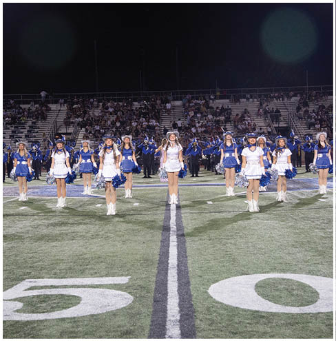 A group of people dressed in blue and white uniforms are standing in formation on a football field at night with a crowd in the bleachers behind them. Description generated by AI