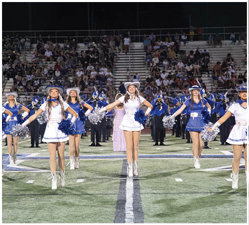 A group of people dressed in coordinated blue and white cheerleading uniforms with hats and pom-poms perform a routine on a football field with a marching band and a crowd in the background. Description generated by AI