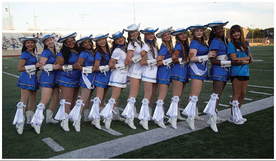 A group of people dressed in blue and white dance team uniforms with hats and boots stand in a line on a football field with their arms linked and legs raised. Description generated by AI