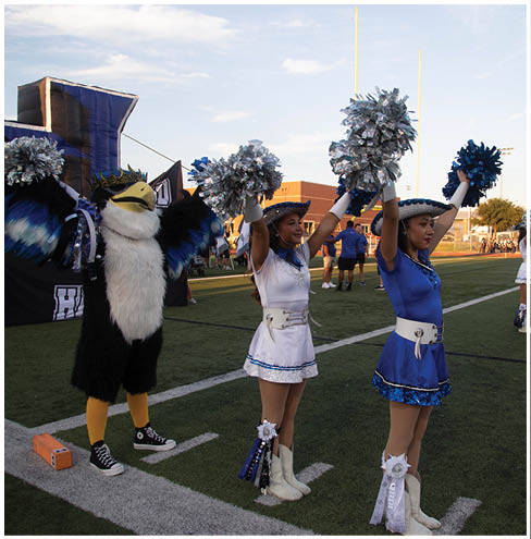 Three cheerleaders in blue and white uniforms and a person in a bird mascot costume are standing on a football field holding pom-poms raised in the air during a daytime event. Description generated by AI